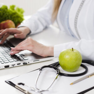 Dietetyk w białym fartuchu pracuje przy laptopie na jasnym biurku, na którym leży stetoskop, zielone jabłko oraz podkładka z długopisem. W tle widoczne są świeże warzywa i owoce. Zdjęcie przedstawia profesjonalne przygotowanie do wywiadu zdrowotnego, który otwiera każdą pierwszą konsultację dietetyczną online.